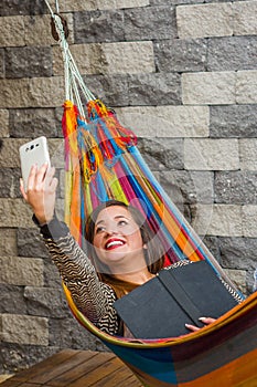 Close up of young beautiful woman relaxing in a hammock and taking a selfie with her tablet while is holding a book with