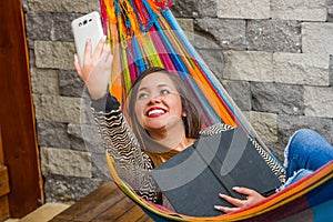 Close up of young beautiful woman relaxing in a hammock and taking a selfie with her tablet while is holding a book with