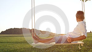 CLOSE UP: Young boy relaxing on a wooden swing gazing at the golden sunset.