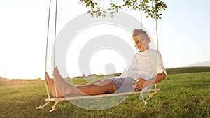 CLOSE UP Young boy relaxing on a wooden swing at sunset,posing for the camera.