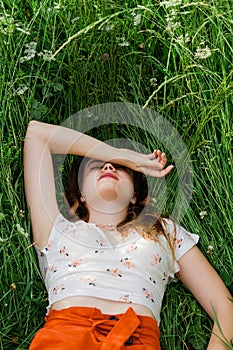 Close up of young brunette girl in white top and red skirt lying down in green meadow field grass in countryside