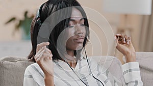 Close-up young carefree woman sitting on sofa in room listening to rhythmic music on headphones enjoying favorite audio