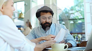 Close up of young Caucasian male and female business partners sitting together at table, pointing on documents in hands