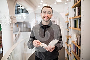 Close-up of young caucasian man with eyeglasses in a bookstore with an opened book in his hands reading something with other books