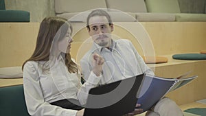 Close-up of young Caucasian man and woman sitting with laptop and documents and talking. Coworkers discussing business