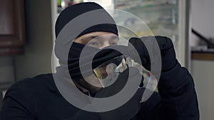 Close-up of young caucasian thief in black clothes raising his balaclava and drinking from the glass. The guy stealing