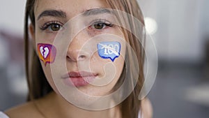 Close-up of young caucasian woman with painted social media icons on her face looking at camera. Addiction to gadgets