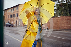 Close-up of a young cheerful woman with a yellow raincoat and umbrella who is in a good mood while walking the city on a rainy day