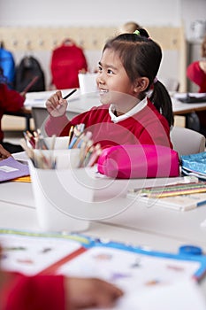 Close up of young Chinese schoolgirl wearing school uniform sitting at a desk in an infant school classroom, selective focus, vert