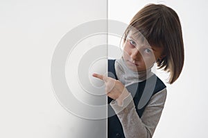 Close up young cute girl looking outside behind white empty banner. She pointing to white wall. Free copy space for design