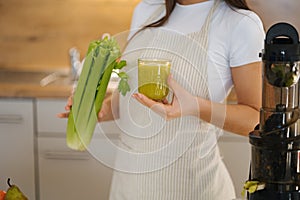 Close-up young female in apron standing in the kitchen. Woman hold glass with green healthy beverage in one hand and