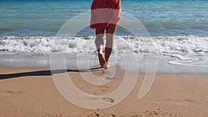 Close up of young female legs walking on sandy beach. Attractive young girl enters into water. Girl in red short dress walking alo
