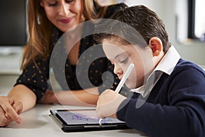 Close up of young female teacher sitting at desk with a Down syndrome schoolboy using a tablet computer in a primary school classr