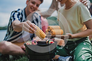 Close-up of young friends putting corn on grill and having barbecue when camping in campground.