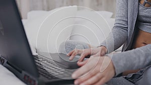 Close up. Young girl in eyeglasses sitting on the bed in a bright room and concentrated on the work in laptop