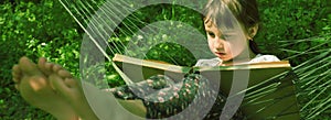 Close up young girl reading book in comfortable hammock against green grass. Pretty female child resting outdoors at  garden.