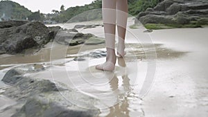 Close up young girl`s legs walking barefoot on the beach with sea water splashing on the sand.