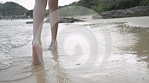 Close up young girl`s legs walking barefoot on the beach with sea water splashing on the sand.