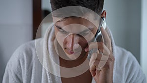 Close-up. A young guy in a white bathrobe talks on a mobile phone while standing in the bathroom.