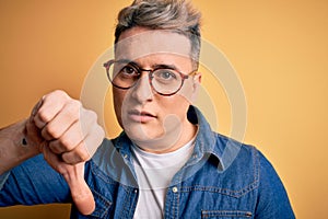 Close up of young handsome modern man wearing glasses and denim jacket over yellow background with angry face, negative sign