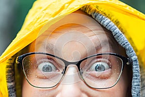 Close-up young happy woman portrait with wide opened amazed eyes wearing glasses with raindrops and bright yellow raincoat hood