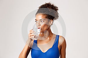 Close-up of young healthy fitness woman drinking glass of water, standing over white background in workout clothing