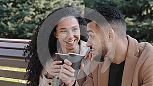 Close up young hispanic couple sitting on bench in autumn city park man and woman drinking coffee husband and wife