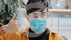 Close-up young indian guy in medical mask posing indoors looking at camera showing thumbs up sign approval agreement