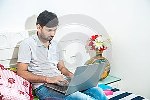 Close up of Young indian man busy working on his laptop doing office work while relaxing on bed in bedroom, freelancer working