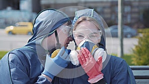 Close-up of young joyful man whispering on ear of pretty woman in chemical suit and respirator. Funny smiling Caucasian