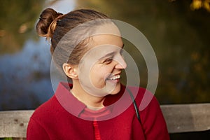 Close up of young lady emotionally laughing, sitting in open air on river bank, side view of happy lady wearing red shirt, has