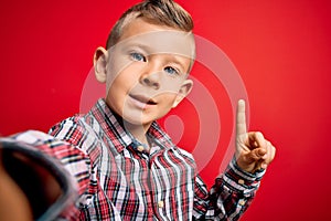 Close up of young little caucasian kid with blue eyes taking a selfie photo over red background surprised with an idea or question