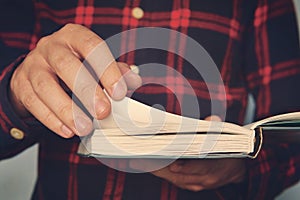 Close up of a young male in a chequered shirt holding the book and turning over the page. Ingusitive man reads the book