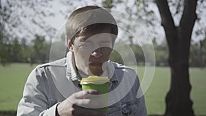 Close-up of young man drinking disgusting coffee outdoors. Portrait of Caucasian guy tasting hot drink and making nasty