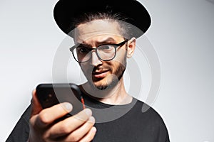 Close-up of young man looking surprised in smartphone, wearing hat and glasses.