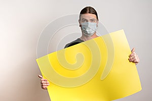 Close up of a young man with protective mask against virus epidemy is holding an empty yellow cardboard against white photo