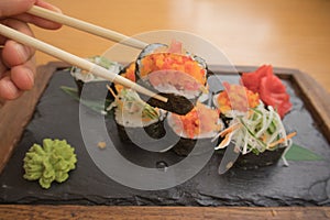 Close up of a young man`s hand holding and eating vegetarian sushi rolls with chopsticks on a wooden tray on a wooden table