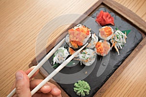 Close up of a young man`s hand holding and eating vegetarian sushi rolls with chopsticks on a wooden tray on a wooden table