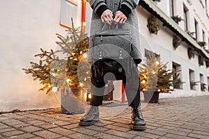 Close-up of a young man standing near the building in a stylish plaid coat in vintage trousers in fashionable boots with a leather