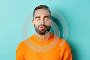 Close-up of young man in sweater pucker lips, close eyes and waiting for kiss, standing over light blue background