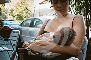 Close up of young mom breastfeeding newborn baby daughter in public outdoors in the street on a bench