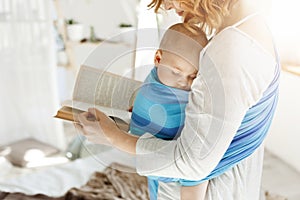 Close up of young mom reading fairy tales for her newborn little son in comfortable light bedroom. Baby falls asleep