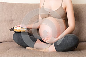 Close up of young pregnant woman enjoys eating tasty cake resting on the sofa at home. Unhealthy diet during pregnancy concept