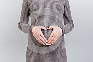 Close up of young pregnant woman in gray dress making heart shape with her hands on the belly at gray background. Love to the