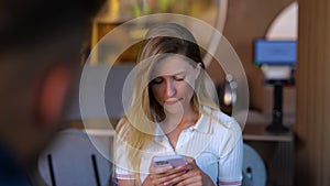 Close up of young pretty Caucasian confused woman texting on smartphone in restaurant. Upset beautiful blonde female