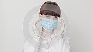 Close-up of young smiling female Caucasian doctor in white coat and protective gloves putting on medical mask and