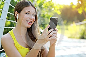 Close up of young smiling woman sitting in the park on summer morning holding smart phone in hands and listening to music through