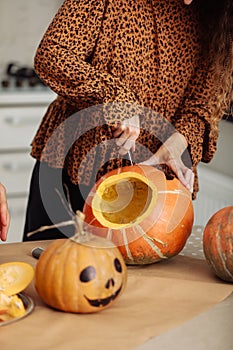 Close up of young woman carving Jack O Lattern from ripe orange pumpkin with knife on her wooden kitchen table. Female