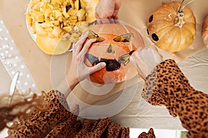 Close up of young woman carving Jack O Lattern from ripe orange pumpkin with knife on her wooden kitchen table. Female