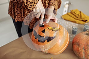 Close up of young woman carving Jack O Lattern from ripe orange pumpkin with knife on her wooden kitchen table. Female